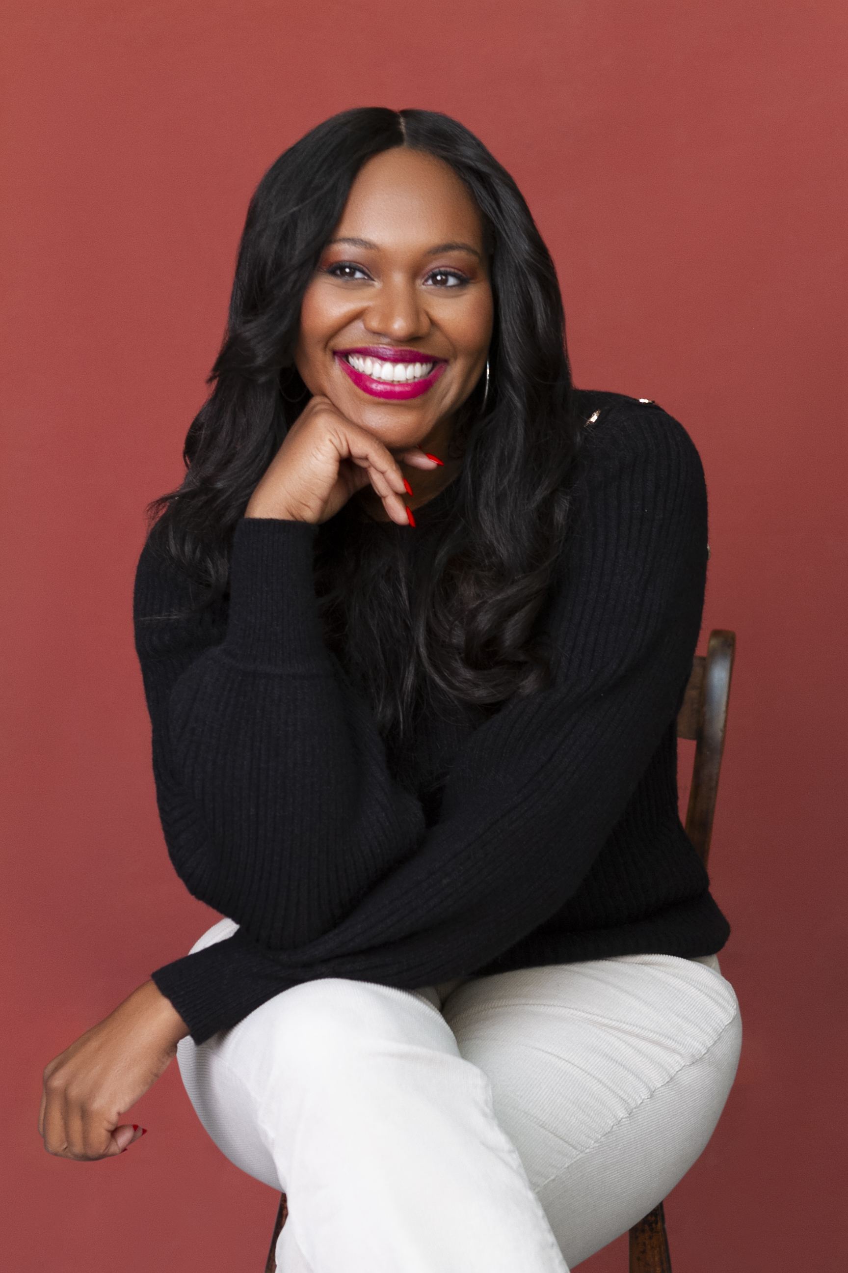 Natalia Anderson wearing sits on chair resting her head on her hand with a smile. Wearing a black jumper and cream trousers